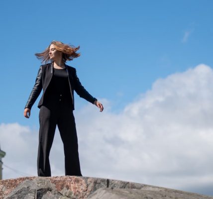 Young woman posing confidently on a rock in a Helsinki park, with a clear blue sky and European architecture in the background.