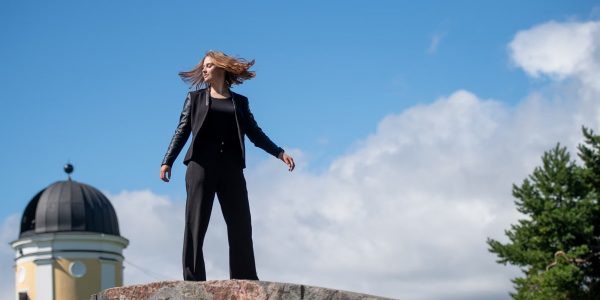 Young woman posing confidently on a rock in a Helsinki park, with a clear blue sky and European architecture in the background.