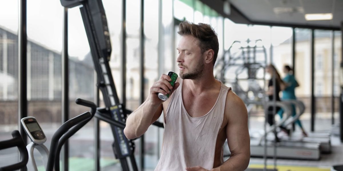 Muscular young sportsman in casual sportswear drinking fresh energy beverage near window while resting after intense workout in modern fitness club