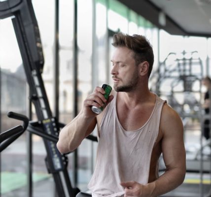 Muscular young sportsman in casual sportswear drinking fresh energy beverage near window while resting after intense workout in modern fitness club