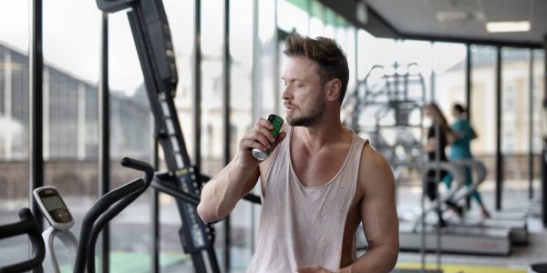 Muscular young sportsman in casual sportswear drinking fresh energy beverage near window while resting after intense workout in modern fitness club